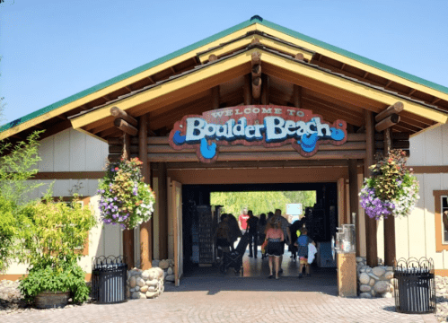 Entrance to Boulder Beach with a wooden sign, colorful flower baskets, and visitors walking inside.